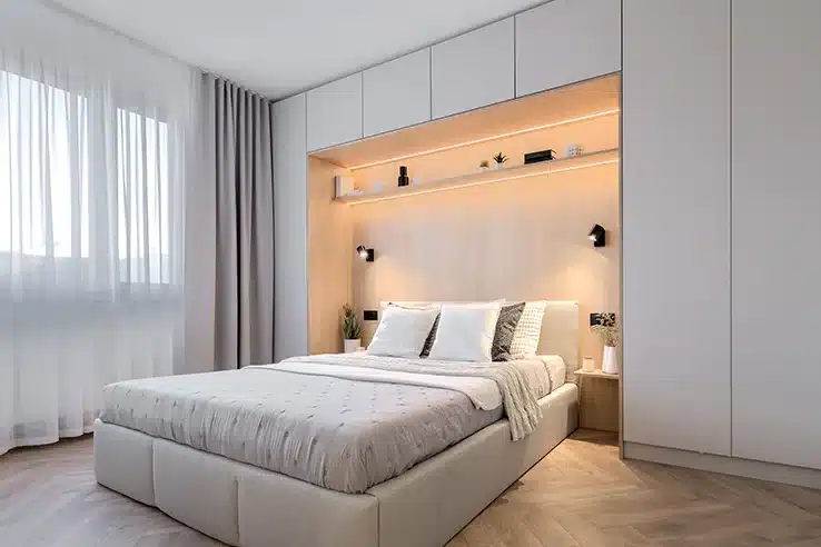 Las pequeñas decisiones que hacen que una vivienda parezca mucho más amplia Dormitorio con armarios empotrados y cabecero de madera con iluminación integrada.